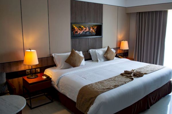 A well-lit hotel room with a large bed, bedside tables with lamps, a wall-mounted fireplace, and a window with curtains drawn to the side.