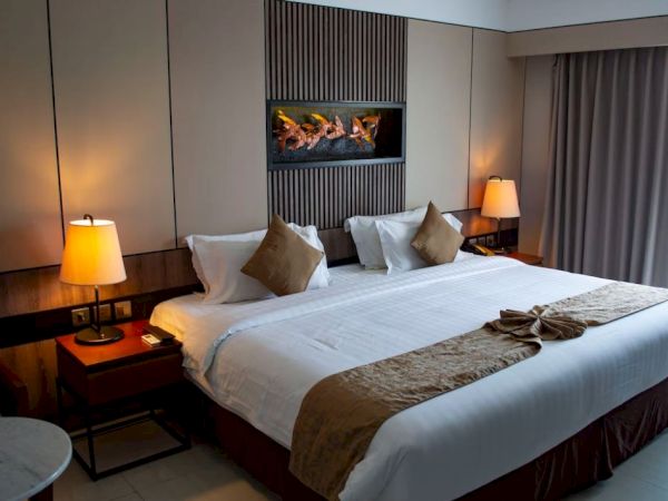 A well-lit hotel room with a large bed, bedside tables with lamps, a wall-mounted fireplace, and a window with curtains drawn to the side.