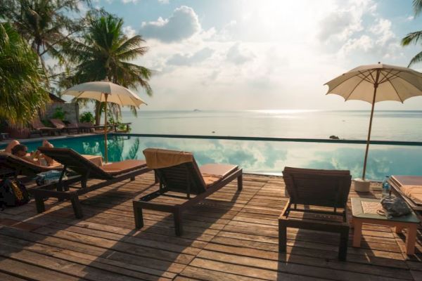A scenic poolside area with lounge chairs, umbrellas, and a serene view of the ocean under a partly cloudy sky. Palms add to the tropical vibe.
