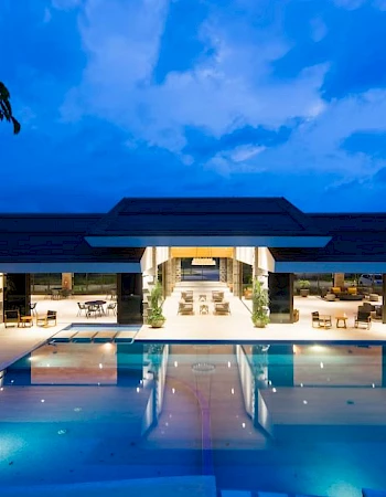 A luxurious open-air pavilion with a large swimming pool illuminated at night, surrounded by lounge chairs and trees, under a vibrant blue sky.