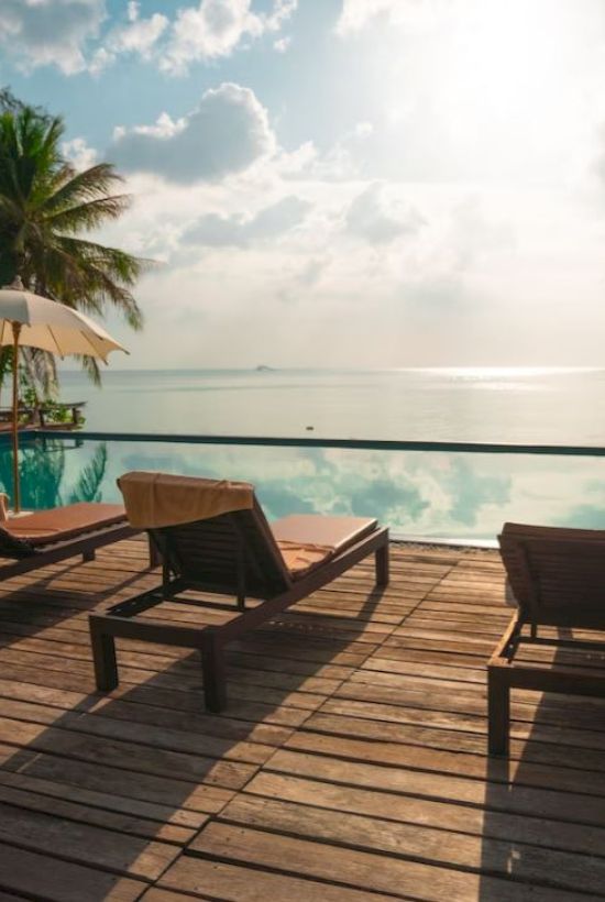 A serene poolside scene with lounge chairs, umbrellas, and a view of the ocean at sunset. Palms and clear skies complete the tropical setting.