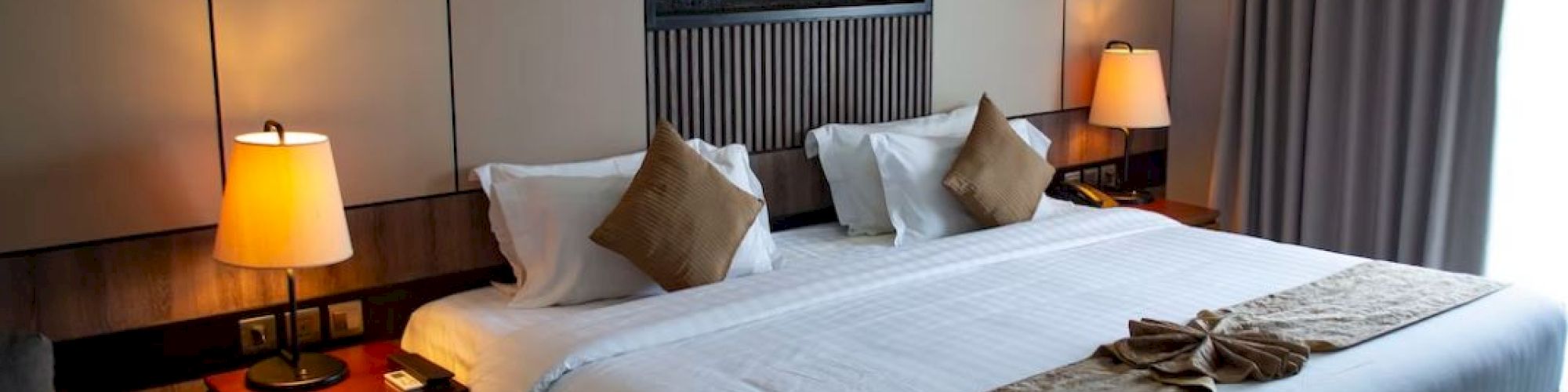 A cozy hotel room with a large bed, pillows, bedside tables, and lamps. There's a decorative wall feature above the bed and closed curtains.