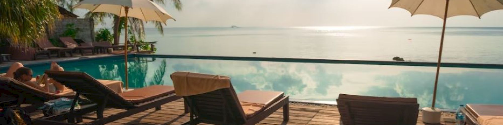 A serene poolside scene with sun loungers and umbrellas overlooks a calm ocean, framed by palm trees and bathed in warm sunlight.