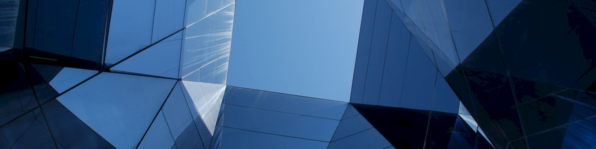 The image shows a view looking up at a geometric pattern created by reflective glass buildings against a blue sky, forming a symmetrical design.