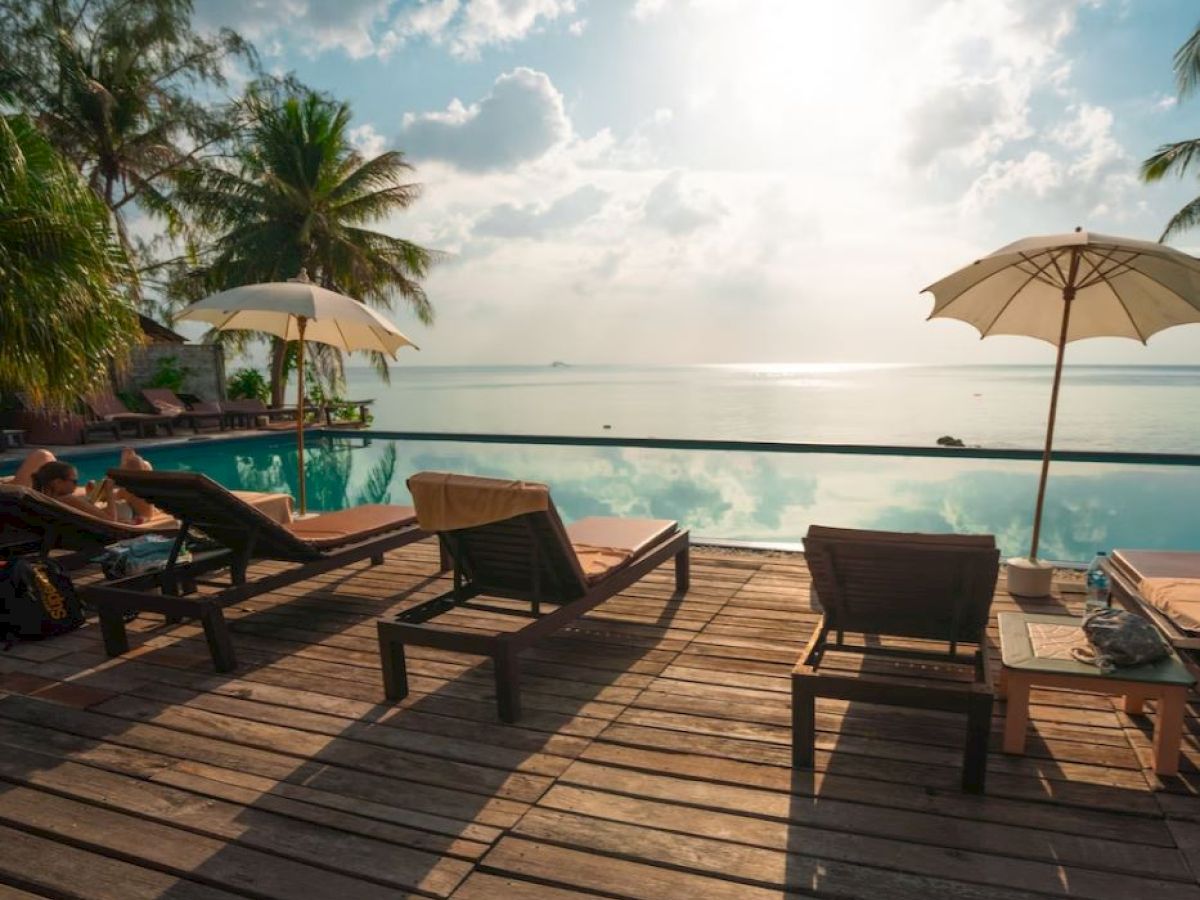 A serene poolside setting with lounge chairs, umbrellas, and palm trees overlooking a calm body of water under a bright, cloudy sky.