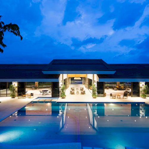 An outdoor pool area is illuminated at night with a modern building in the background, featuring large open spaces and seating areas.