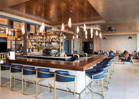 The image shows a modern bar with a wooden counter, blue barstools, hanging lights, and a stocked liquor shelf, set in a spacious seating area.