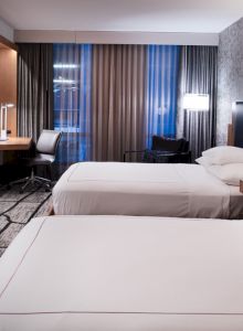 A modern hotel room with two beds, a wall-mounted TV, a desk with a chair, and stylish decor, including patterned wallpaper and curtains, is shown.