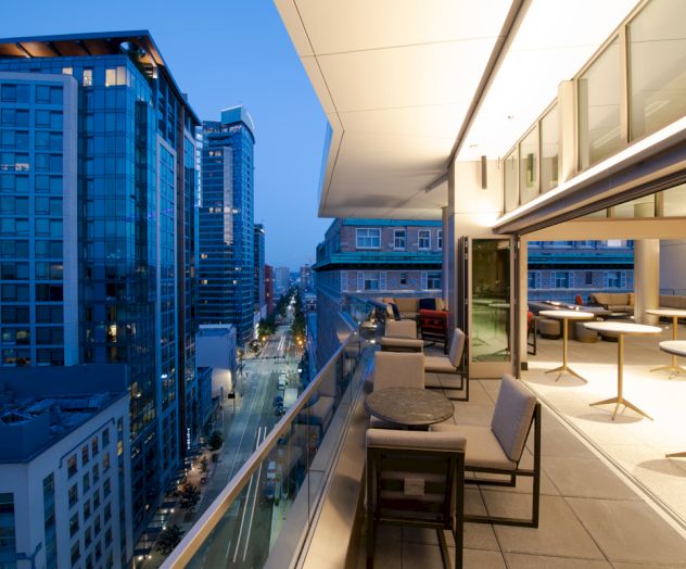 A modern rooftop terrace with seating overlooks a city street lined with tall buildings during twilight, offering a panoramic urban view.