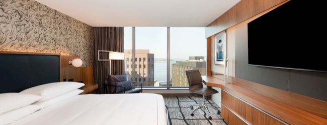 A modern hotel room with a large bed, a desk with a chair, a flat-screen TV, and a window offering a cityscape view.