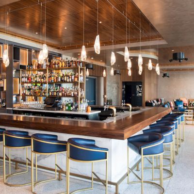 A modern bar with sleek blue stools, a well-stocked liquor shelf, and chic pendant lights, with a seating area in the background.