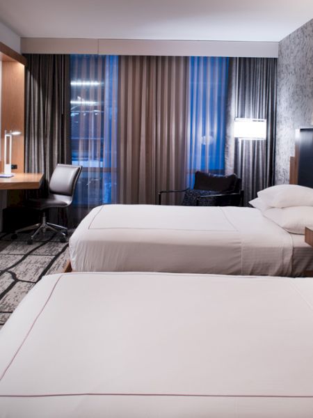 A modern hotel room with two neatly made beds, a wall-mounted TV, a work desk and chair near the window, and stylish decor.