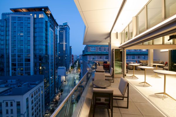 A modern high-rise balcony view at dusk, featuring outdoor seating and tables, overlooking a city street lined with tall buildings and lights.
