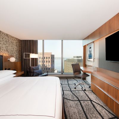 A modern hotel room with a large bed, flat-screen TV, desk, and large window offering a city view.