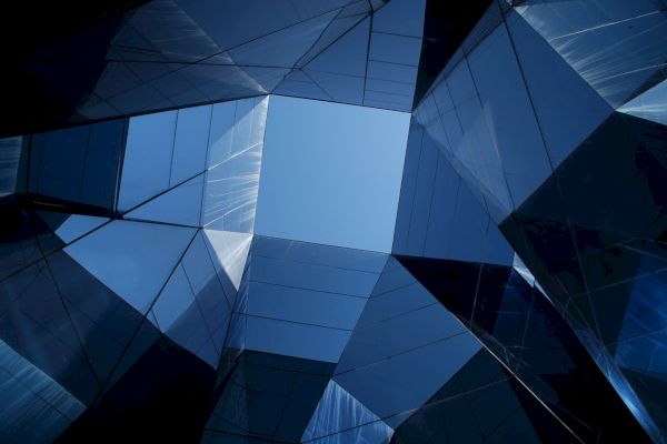 The image shows a geometric arrangement of mirrored glass panes in a structure, creating a reflective pattern with a clear blue sky visible in the center.