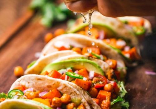 The image shows a hand squeezing lime over several tacos filled with chickpeas, vegetables, avocado, and cilantro, arranged on a wooden surface.