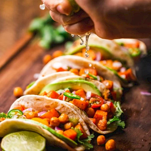 The image shows a hand squeezing lime over several tacos filled with chickpeas, vegetables, avocado, and cilantro, arranged on a wooden surface.