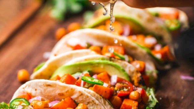 The image shows a hand squeezing lime over several tacos filled with chickpeas, vegetables, avocado, and cilantro, arranged on a wooden surface.