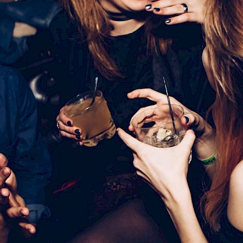People are socializing in a dimly lit setting, holding drinks and smiling, suggesting they are having a fun and lively time together.