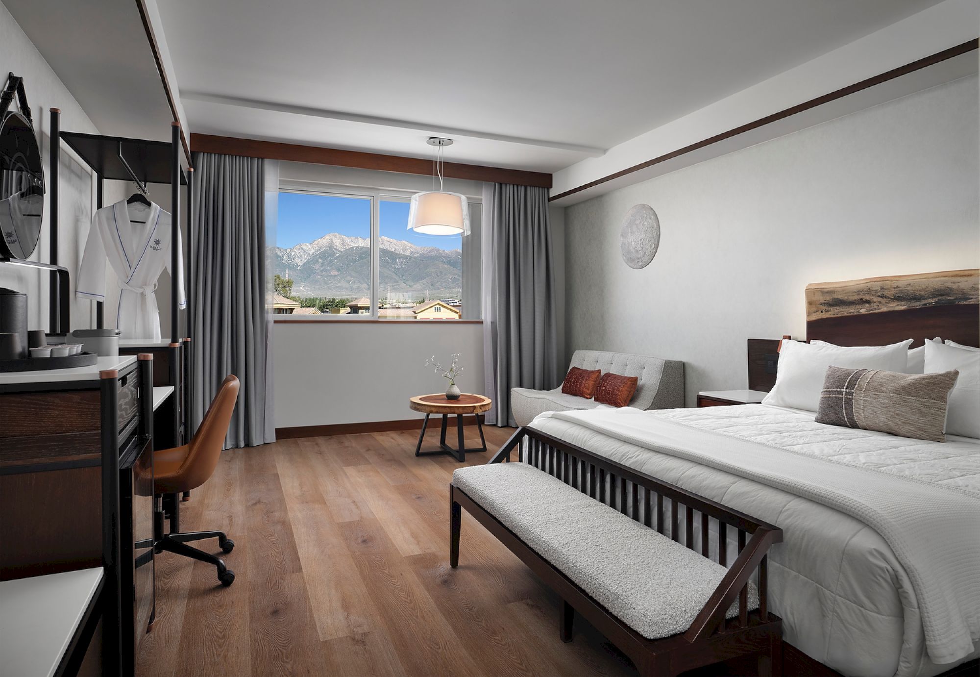 A modern hotel room with a double bed, lounge chair, desk, and window view of mountains; decor includes robes and a round wall artwork.
