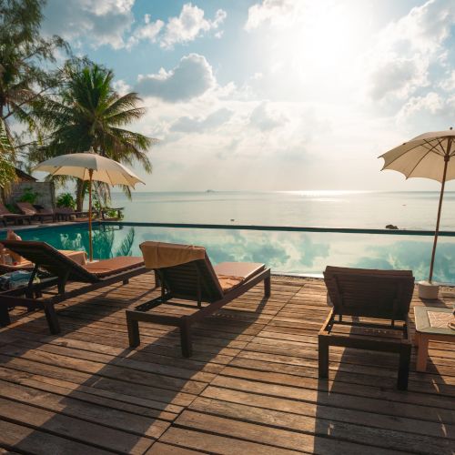 A serene tropical setting featuring lounge chairs and umbrellas on a wooden deck, overlooking an infinity pool and the ocean at sunset.