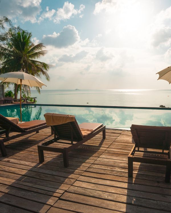 A serene tropical setting featuring lounge chairs and umbrellas on a wooden deck, overlooking an infinity pool and the ocean at sunset.