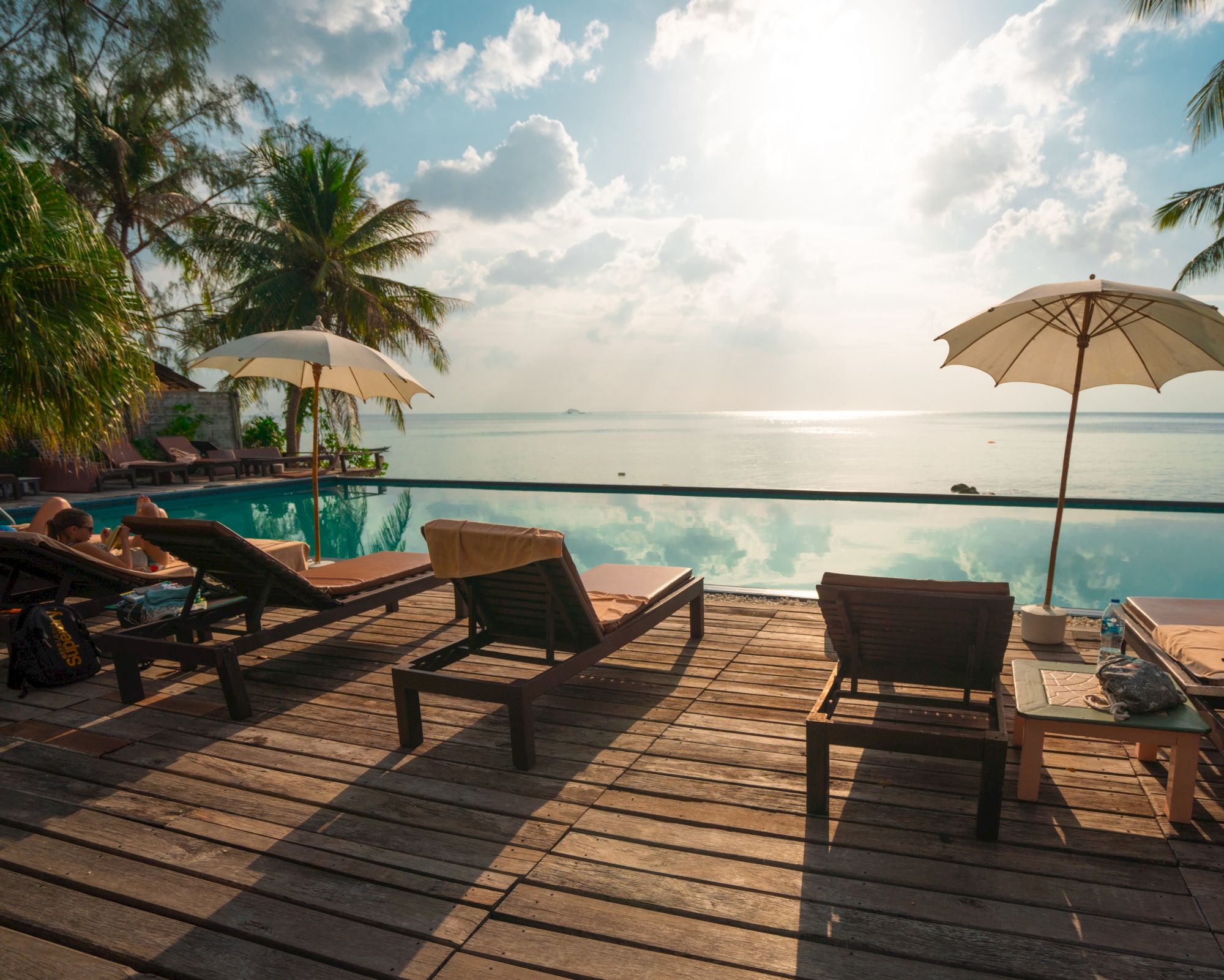 Wooden deck with sun loungers and umbrellas facing an infinity pool, overlooking a calm sea with a bright sky.