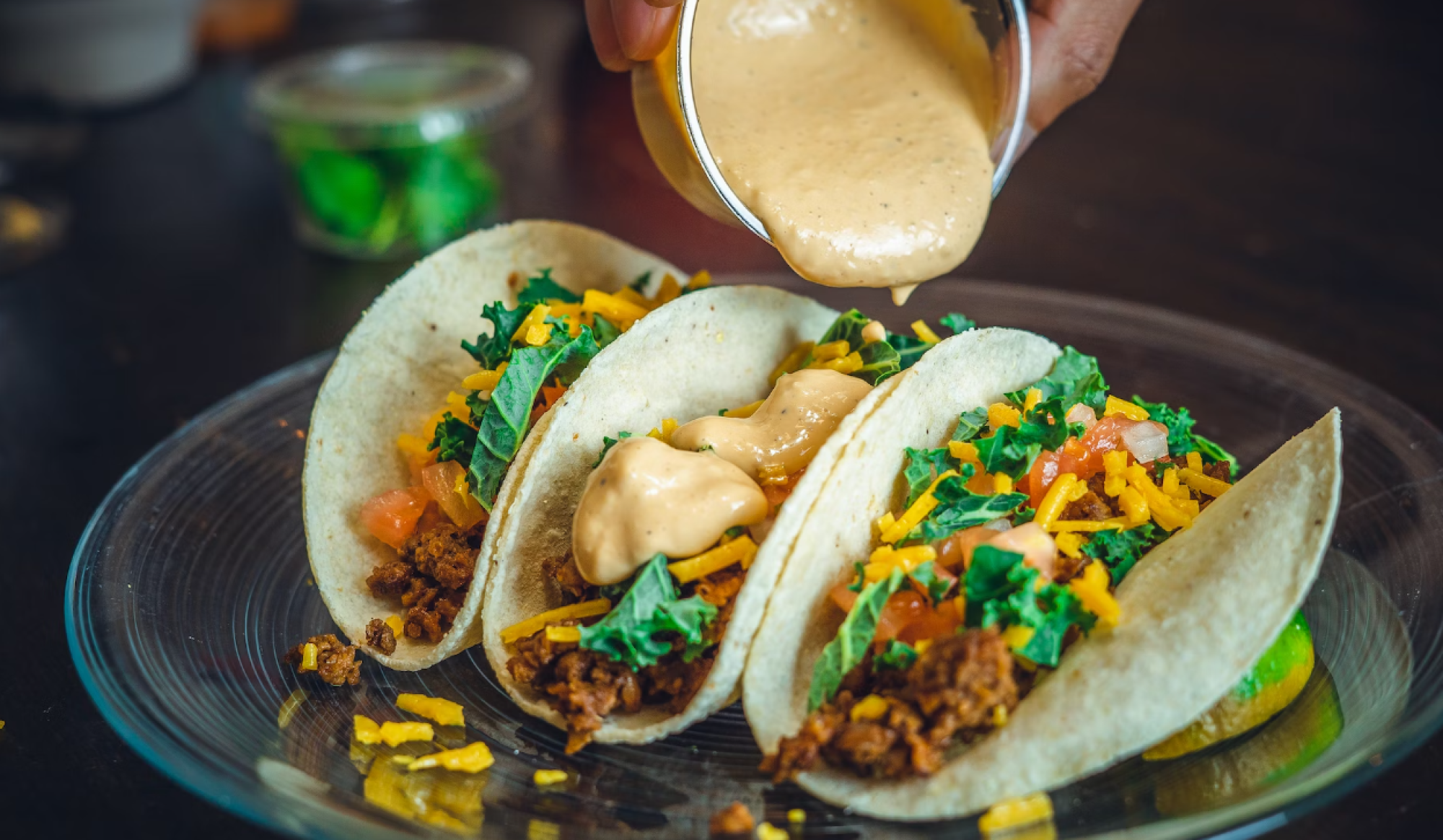Three tacos filled with ground meat, vegetables, and cheese, being topped with a creamy sauce on a clear plate.