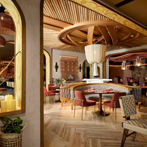 A stylish, modern restaurant interior with wooden decor, woven chairs, plants, and artistic light fixtures, creating a cozy ambiance.