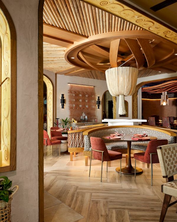 A stylish, modern restaurant interior with wooden decor, woven chairs, plants, and artistic light fixtures, creating a cozy ambiance.