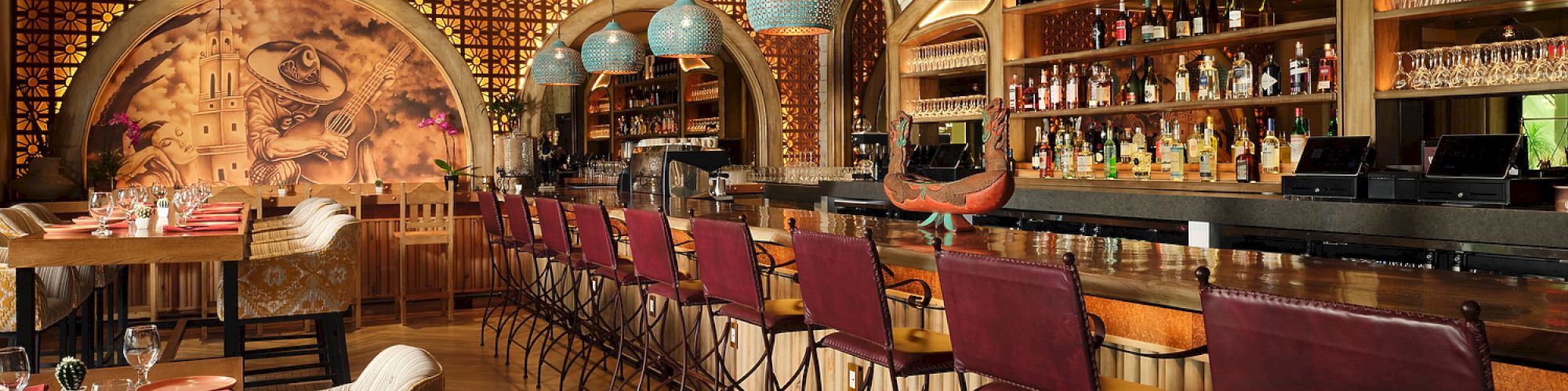 A stylish bar and dining area with red chairs, hanging blue lamps, a mural, and stocked shelves of bottles create a cozy atmosphere.