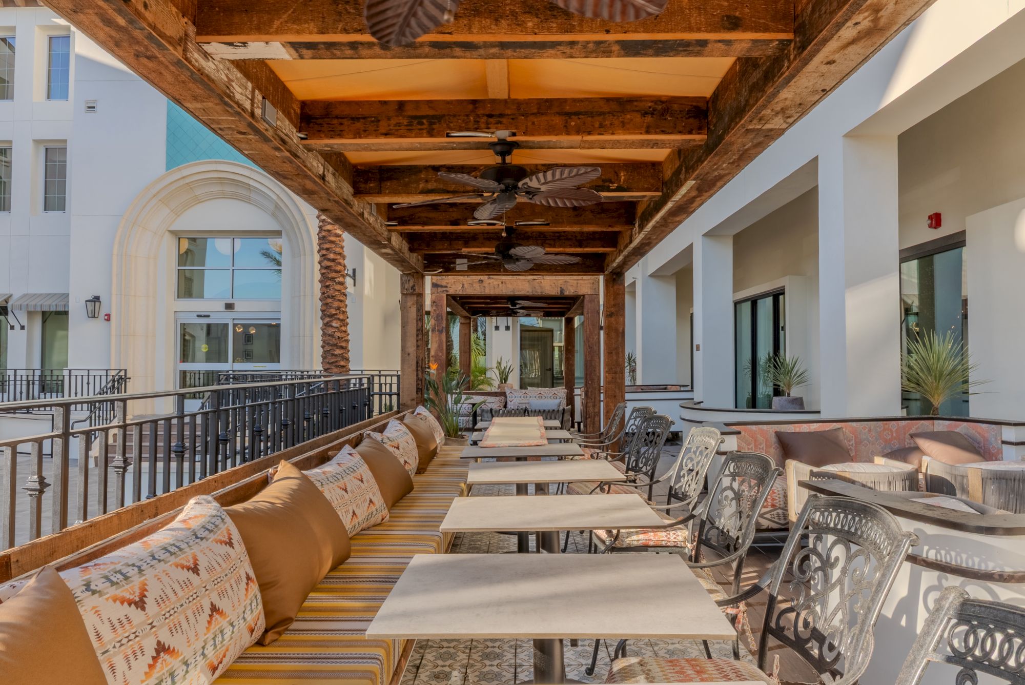 An outdoor seating area with cushioned benches, tables, and potted plants under a wooden pergola with ceiling fans.