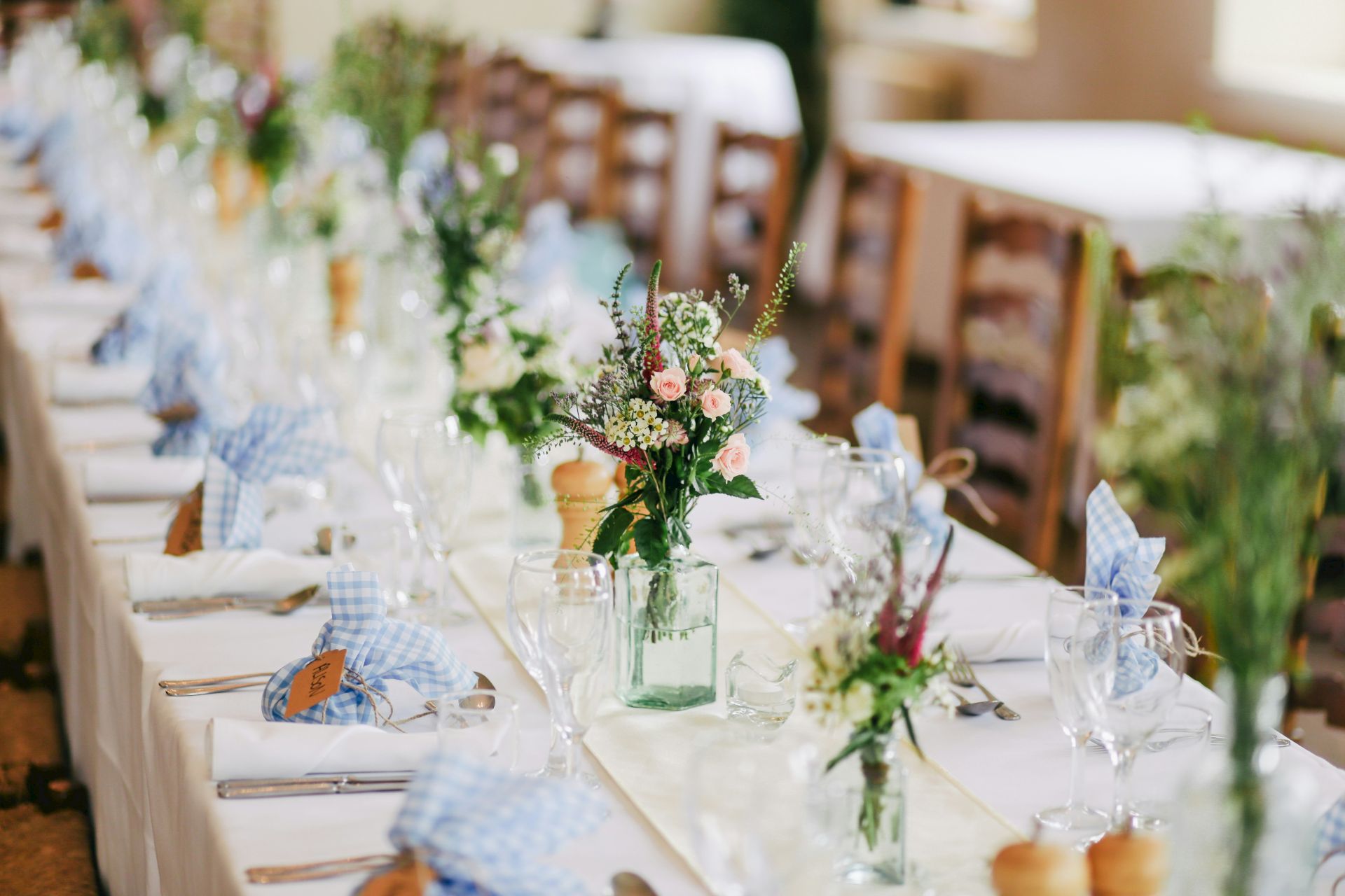 A long table is elegantly set for a formal event, adorned with flowers, glassware, folded napkins, and cutlery on a white tablecloth.