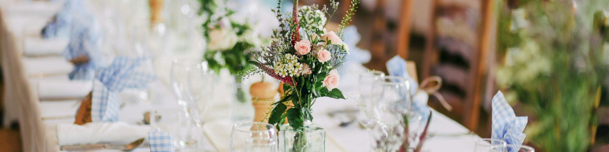 A long table is elegantly set for a formal event, adorned with flowers, glassware, folded napkins, and cutlery on a white tablecloth.