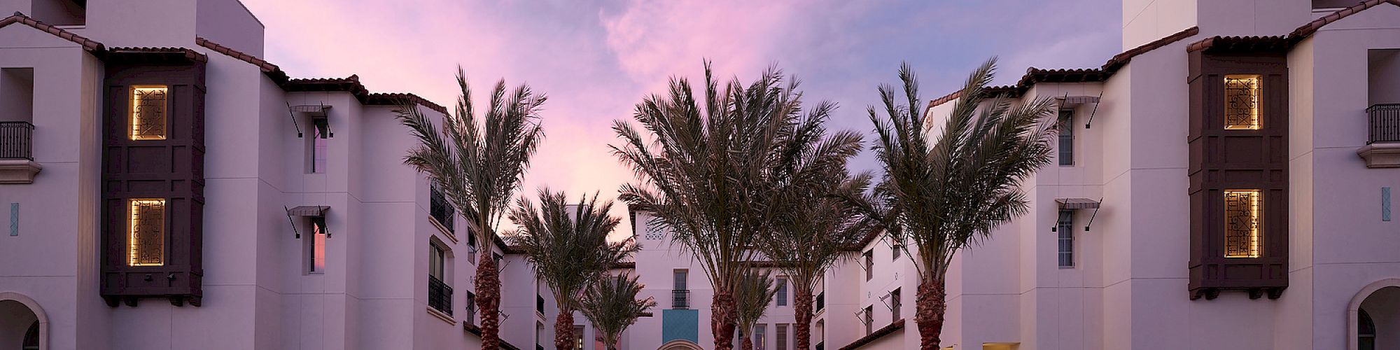 A modern building with palm trees in front, under a pinkish twilight sky, with a symmetrical facade and lit windows.