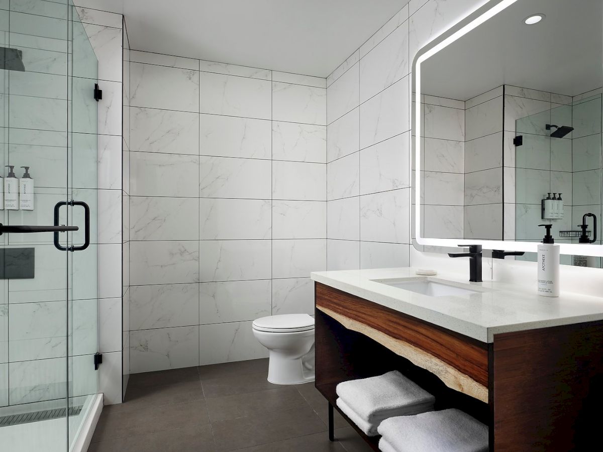 A modern bathroom with a glass shower, illuminated mirror, sink with faucet, and wooden vanity with towels underneath.