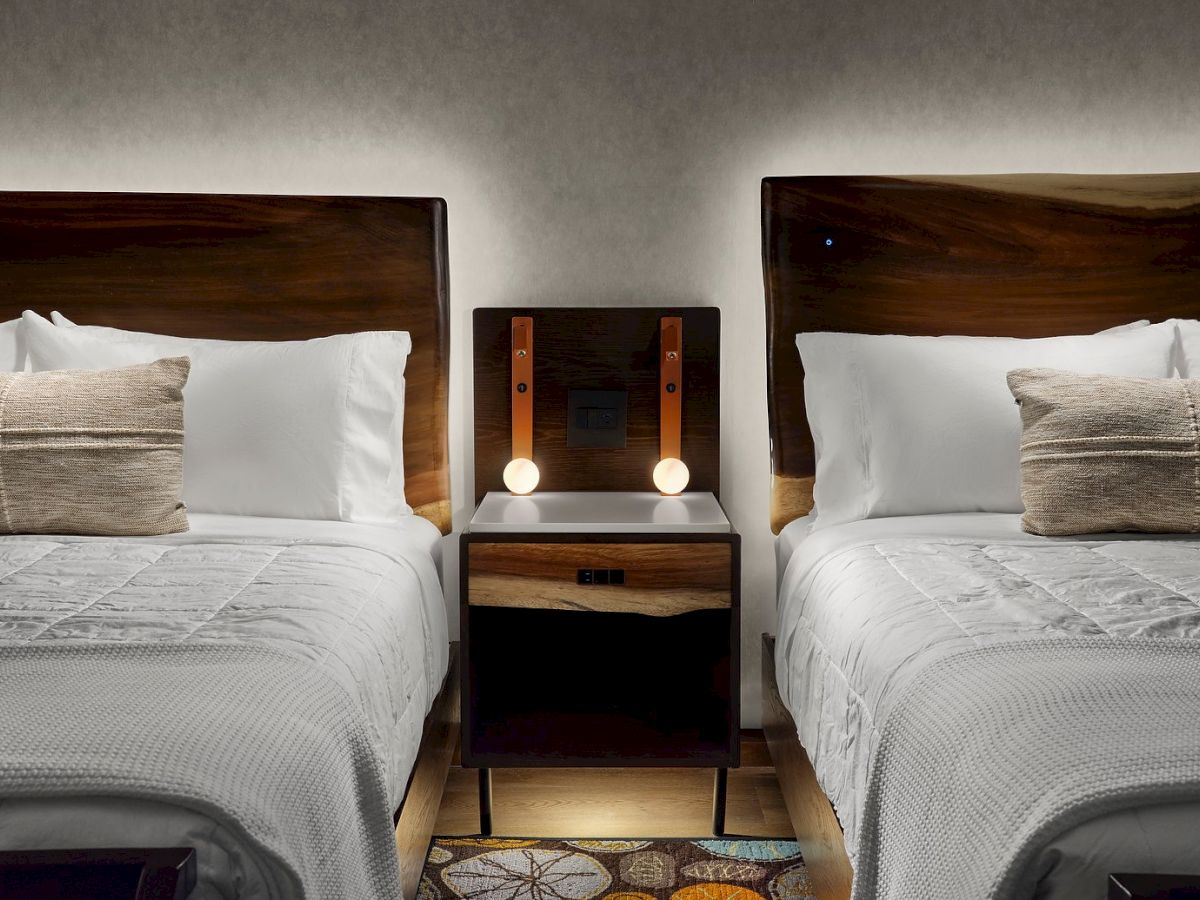 Two beds with wooden headboards and white bedding, separated by a nightstand with two round lights, in an elegantly decorated room.