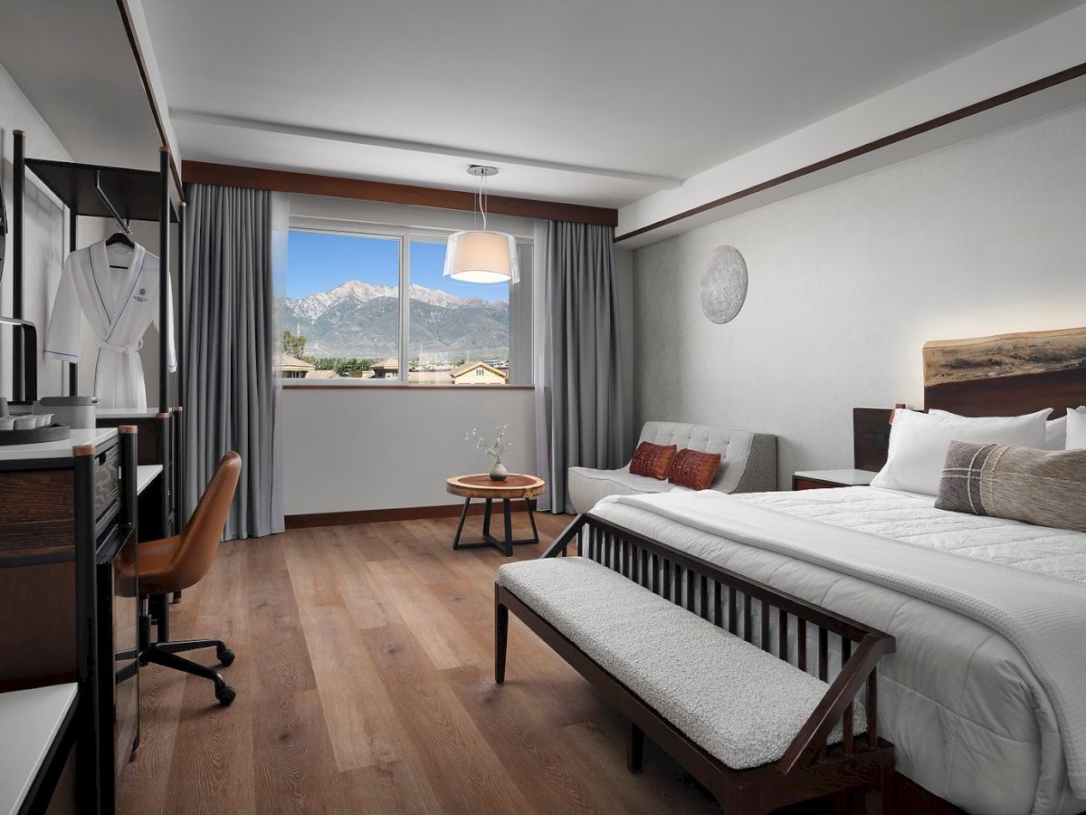 A modern hotel room with a mountain view. It features a bed, sofa, desk, wardrobe, and window showing picturesque scenery.