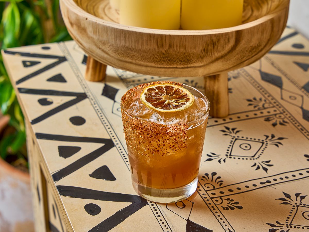 A glass with a drink garnished with a dried citrus slice, on a patterned table next to a wooden bowl with candles.