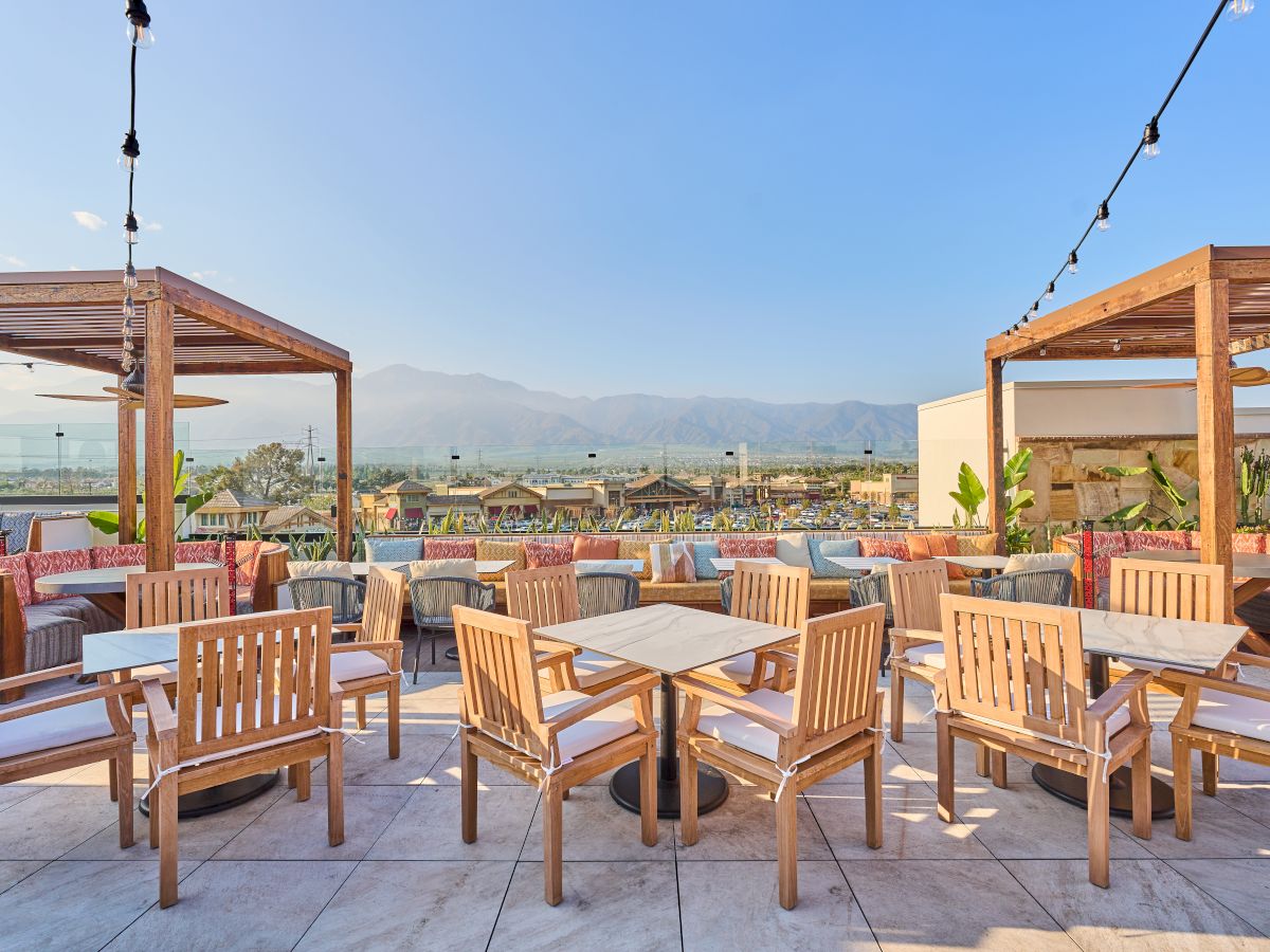 Outdoor seating area with wooden tables and chairs beneath pergolas, overlooking a scenic view with mountains and clear skies in the background.