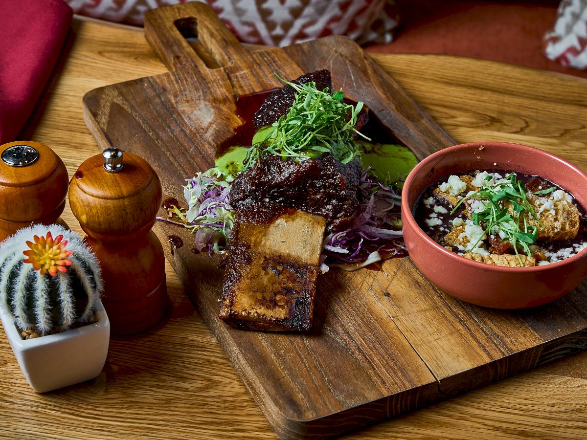 A wooden board with seasoned ribs, greens, and a side bowl of mixed grains. Nearby are salt, pepper shakers, and a small cactus.