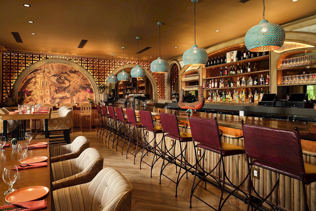A stylish bar with red chairs, ornate hanging lights, colorful mural, and well-stocked shelves, features a cozy and inviting atmosphere.