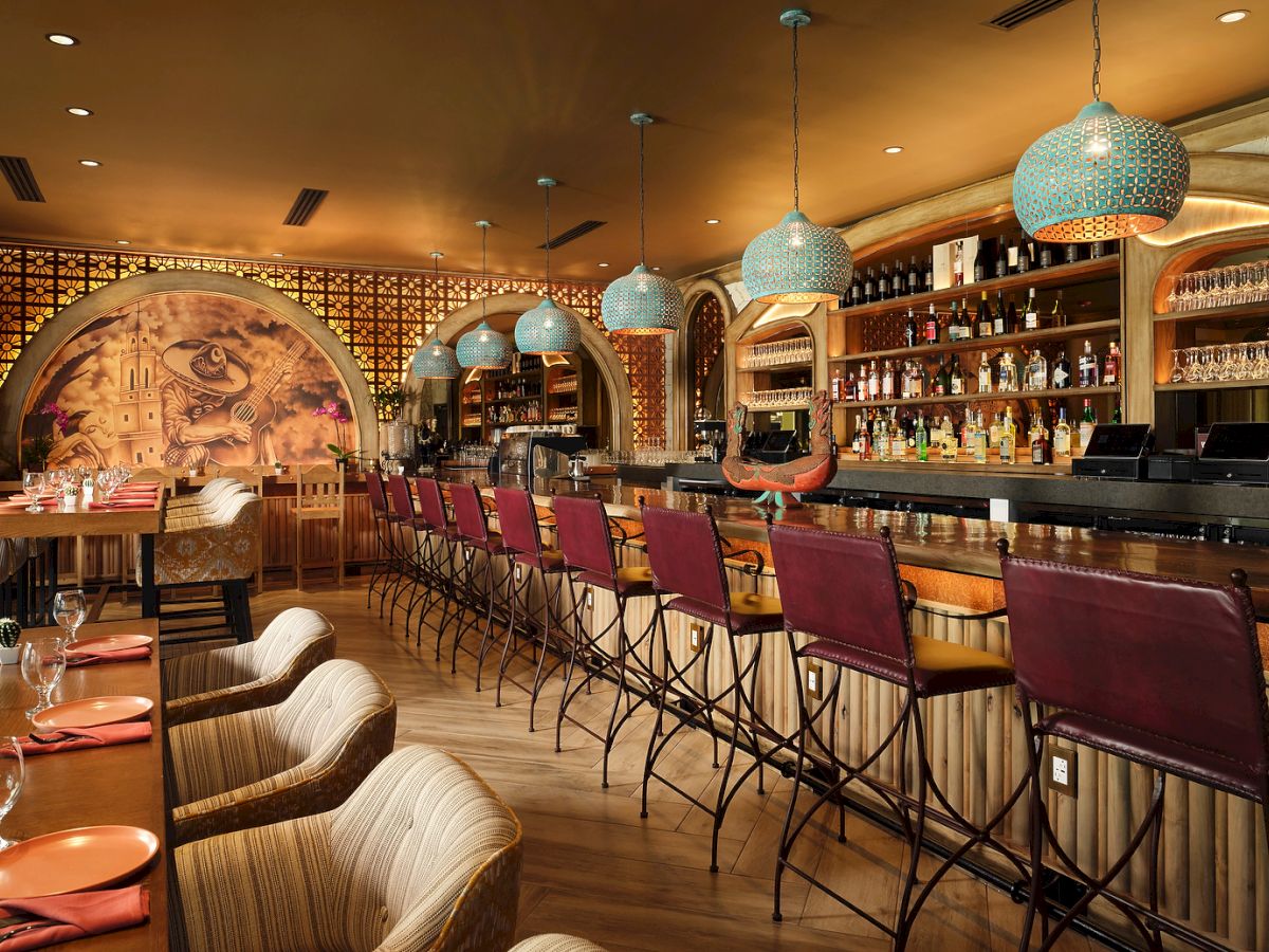 A stylish bar with red chairs, ornate hanging lights, colorful mural, and well-stocked shelves, features a cozy and inviting atmosphere.