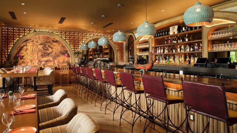 A stylish bar with red chairs, ornate hanging lights, colorful mural, and well-stocked shelves, features a cozy and inviting atmosphere.