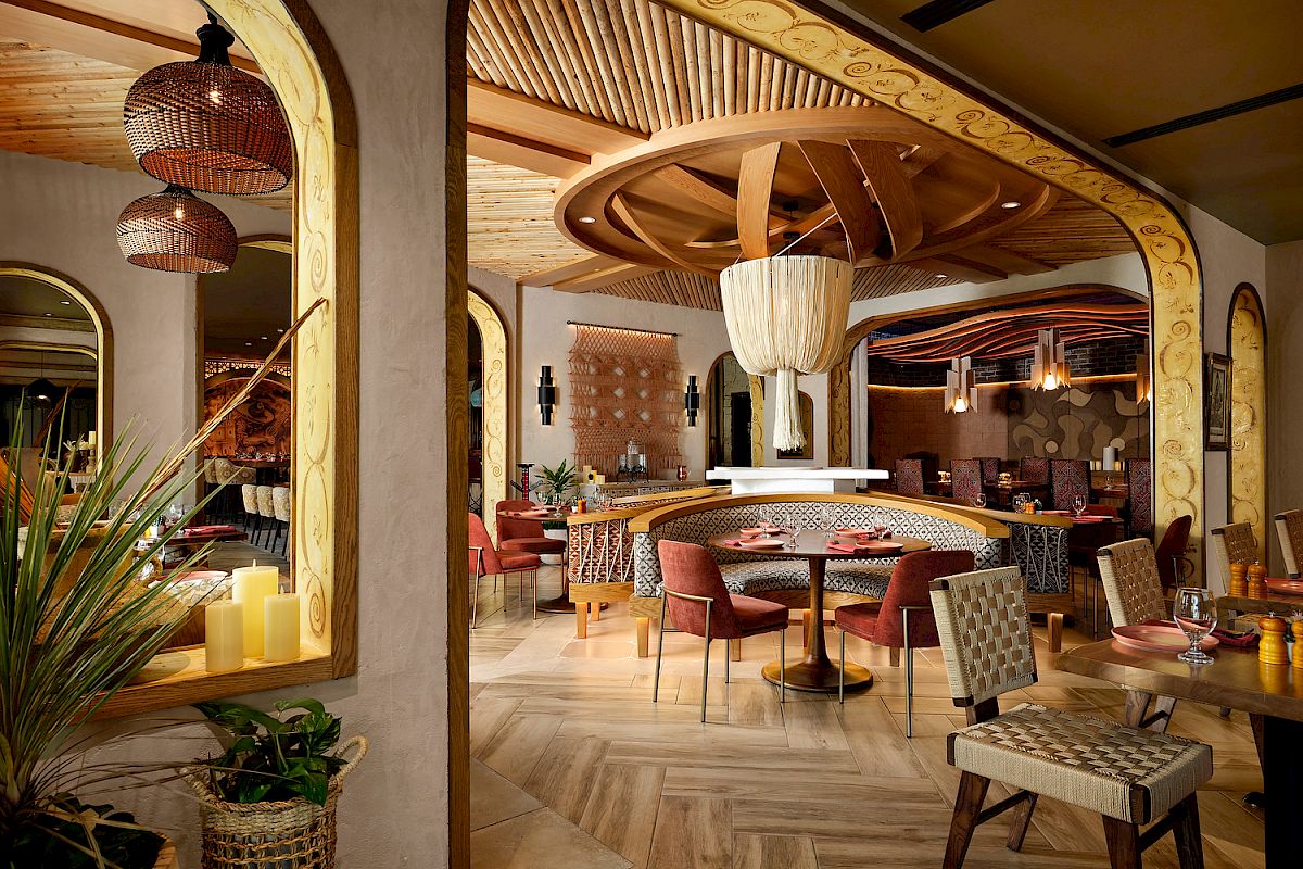 The image shows a stylish restaurant interior with wooden accents, woven chairs, cylindrical lights, and a cozy, inviting atmosphere.
