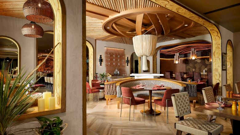The image shows a stylish restaurant interior with wooden accents, woven chairs, cylindrical lights, and a cozy, inviting atmosphere.