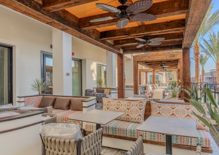 Stylish outdoor patio with patterned sofas, tables, ceiling fans, wooden beams, and palm trees in the background.