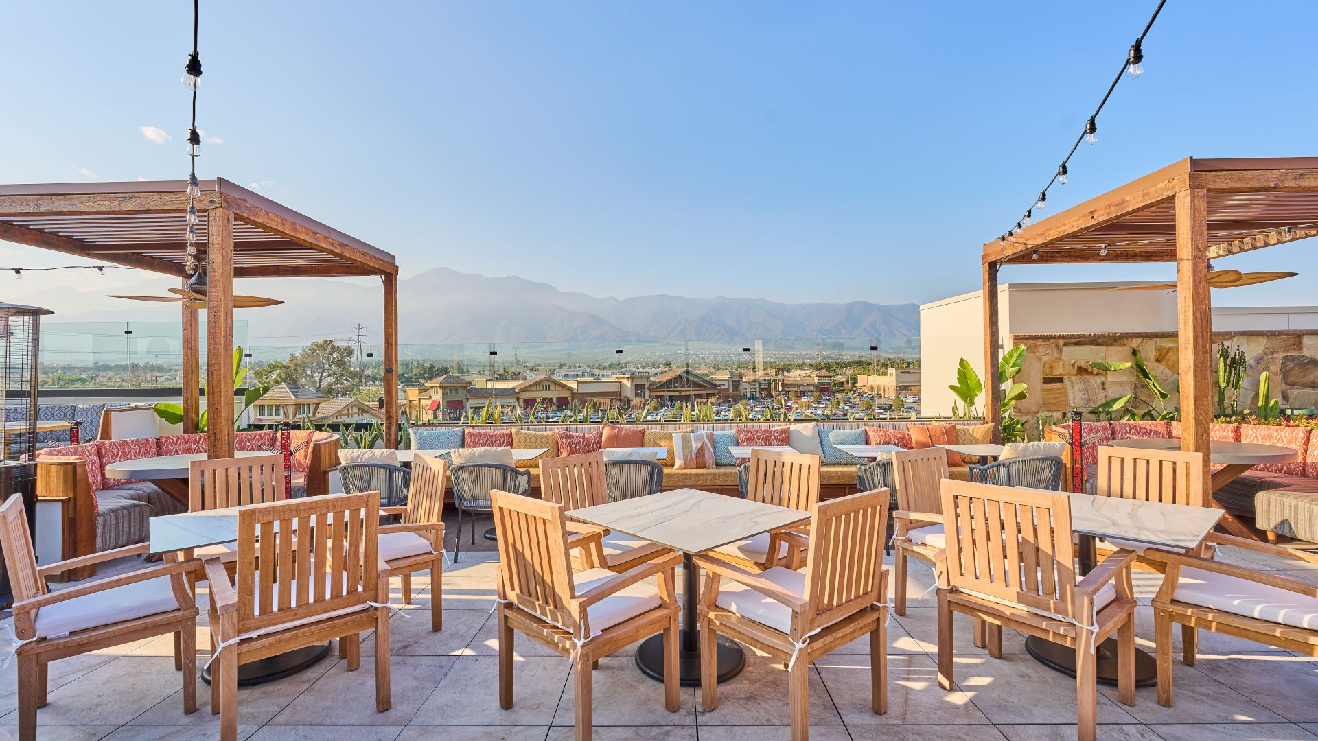 Outdoor seating area with wooden tables and chairs, shaded by pergolas, surrounded by string lights and mountain views in the background.