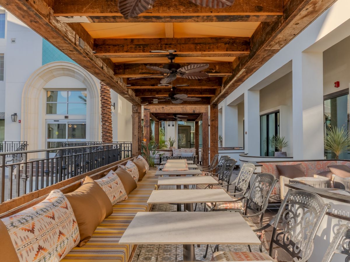 Outdoor seating with patterned cushions, metal chairs, and overhead fans under a wooden pergola. Building and potted plants in the background.
