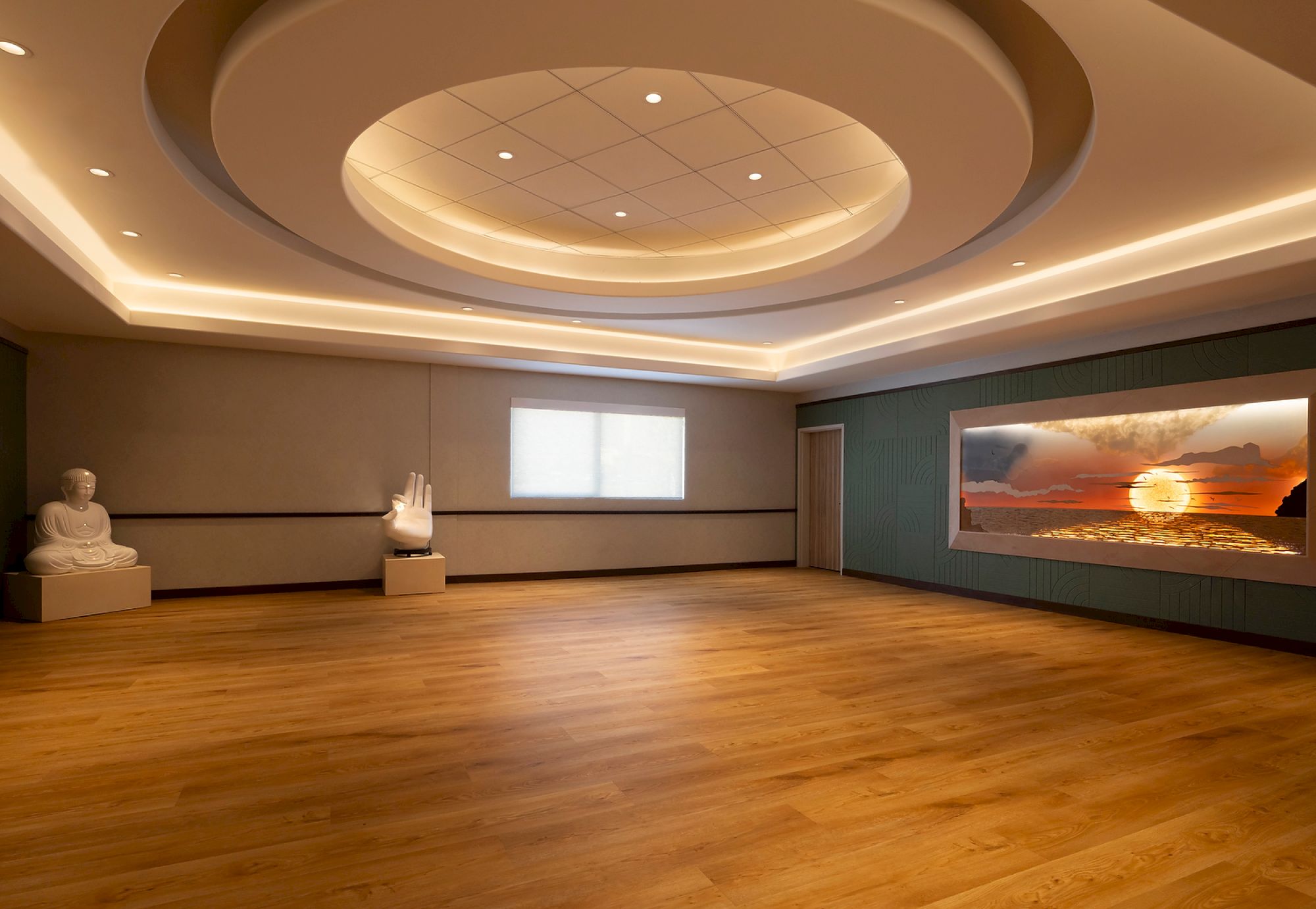 A serene room with wooden flooring, a Buddha statue, hand sculpture, and a large window showing a sunset over water.